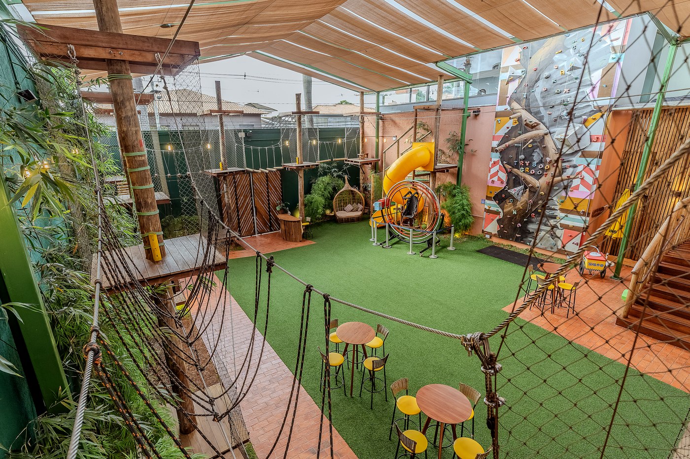 Área externa da TRYBOO, cercada de verde, com arvorismo, escorregador, orbital 360° e parede de escalada