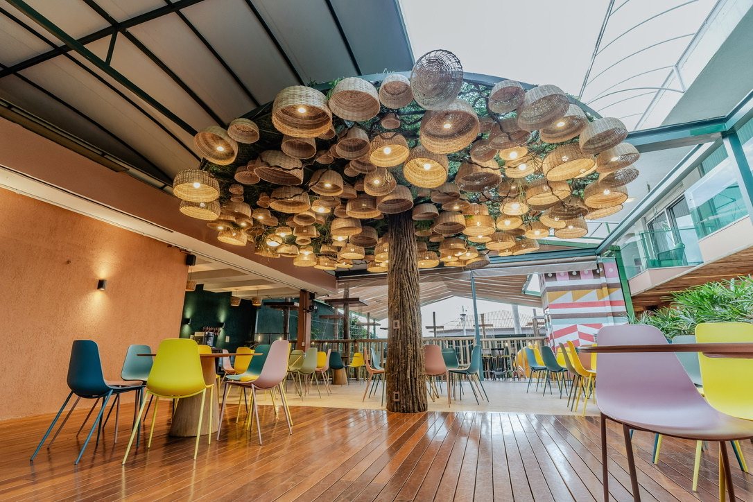 Salão dos adultos da TRYBOO, com árvore de luminárias de palha ao centro, piso de madeira e cadeiras coloridas